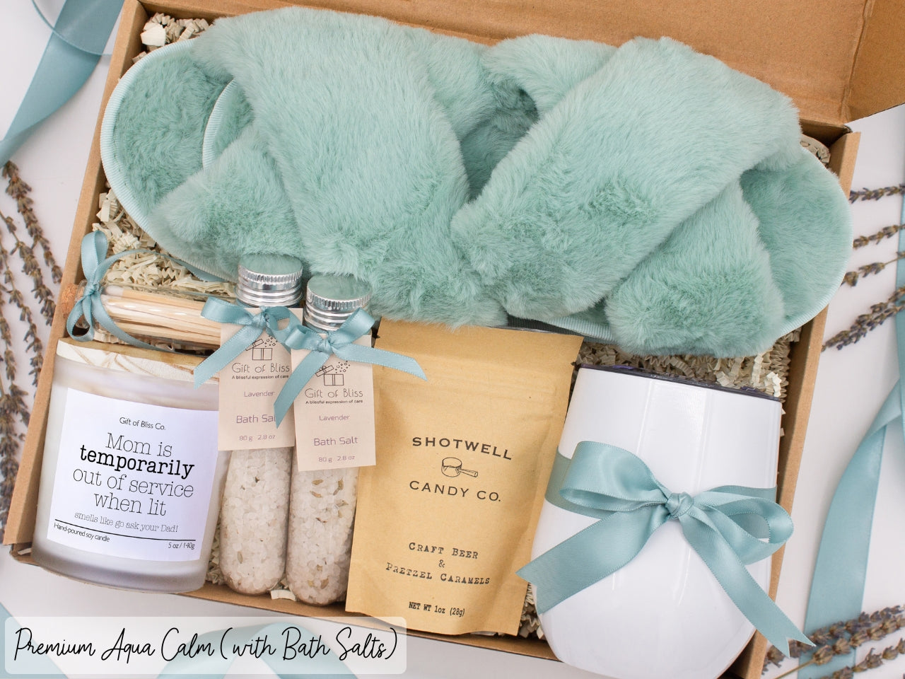 Luxury gift box for her. Featuring a stainless steel tumbler, an aromatherapy candle with mom-themed label and a charming match jar, therapeutic bath salts, plush open-toe slippers, and gourmet caramel treats. All items are beautifully arranged for a pampering experience.