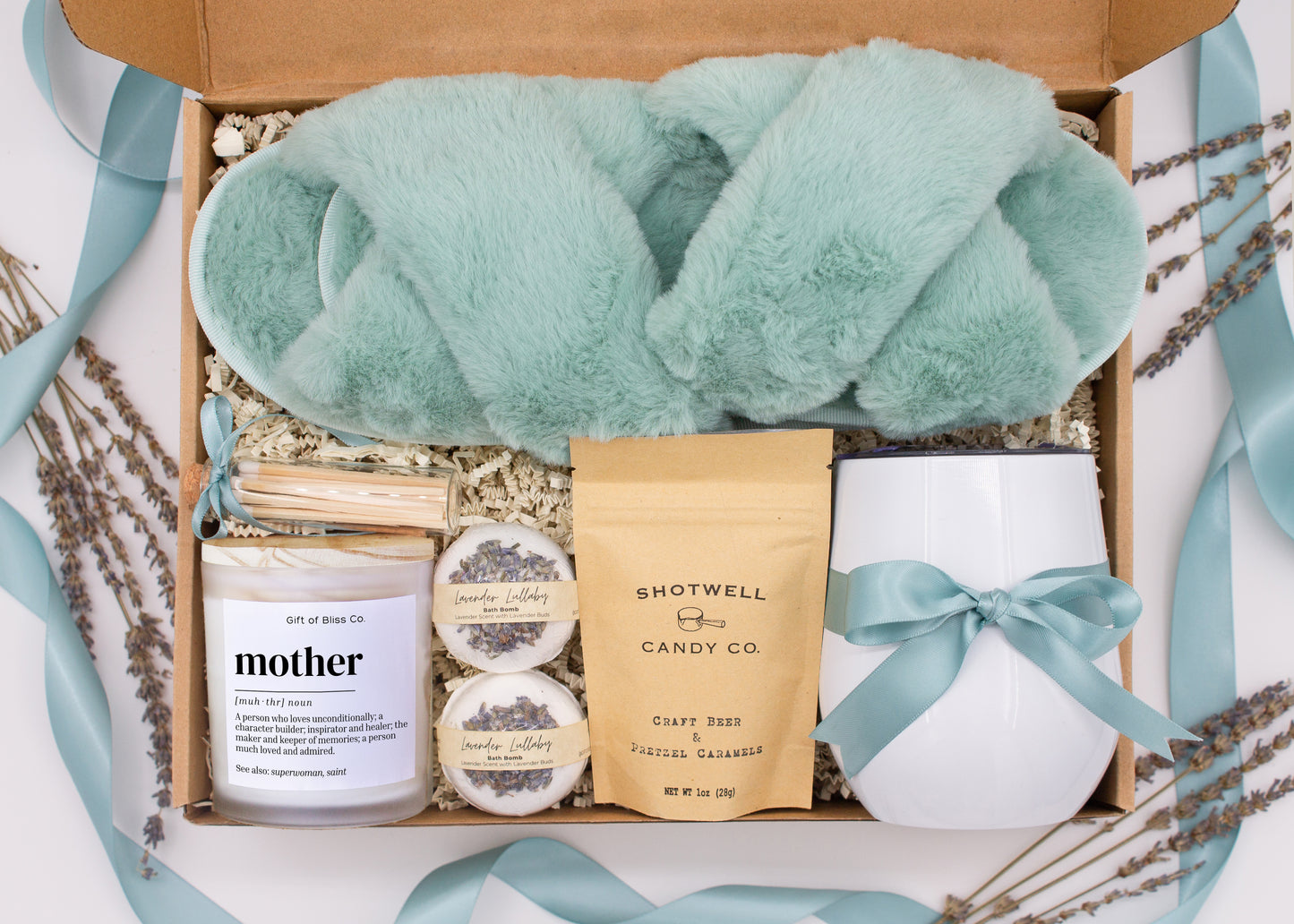 A luxury spa gift box for mom featuring a stainless steel tumbler, an aromatherapy candle with matches, therapeutic bath bombs, plush open-toe slippers, and gourmet caramel treats. All items are beautifully arranged for a pampering experience.