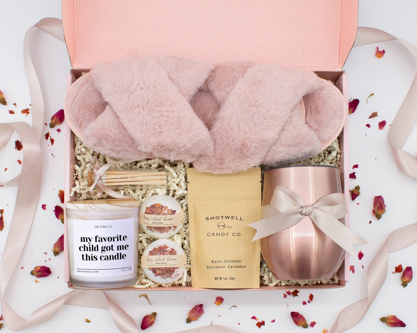 A luxury spa gift box for mom featuring a stainless steel tumbler, an aromatherapy candle with matches, therapeutic bath bombs, plush open-toe slippers, and gourmet caramel treats. All items are beautifully arranged for a pampering experience.