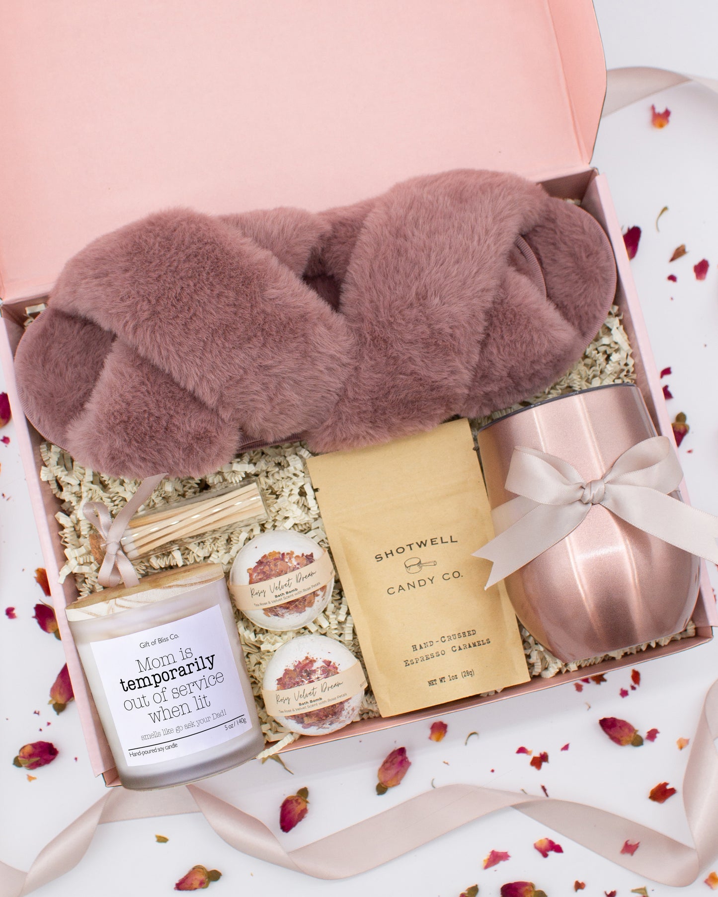 A luxury spa gift box for mom featuring a stainless steel tumbler, an aromatherapy candle with matches, therapeutic bath bombs, plush open-toe slippers, and gourmet caramel treats. All items are beautifully arranged for a pampering experience.