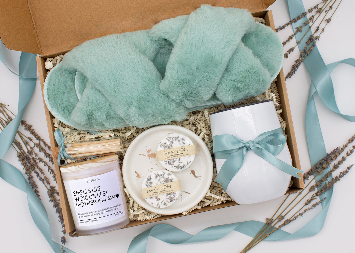 A luxury spa gift box for mom featuring a stainless steel tumbler, an aromatherapy candle with matches, therapeutic shower steamers, plush open-toe slippers, and gourmet caramel treats. All items are beautifully arranged for a pampering experience.