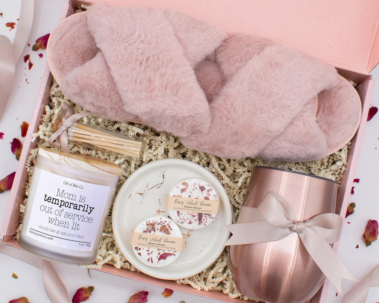 A luxury spa gift box for mom featuring a stainless steel tumbler, an aromatherapy candle with matches, therapeutic shower steamers, plush open-toe slippers, and gourmet caramel treats. All items are beautifully arranged for a pampering experience.