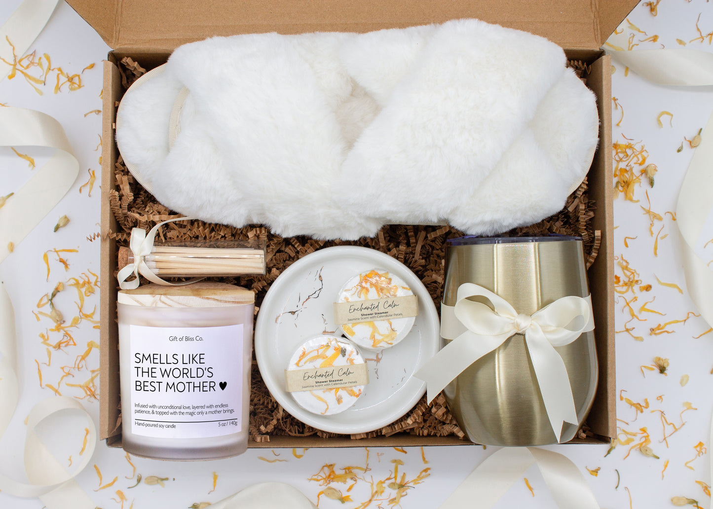A luxury spa gift box for mom featuring a stainless steel tumbler, an aromatherapy candle with matches, therapeutic shower steamers, plush open-toe slippers, and gourmet caramel treats. All items are beautifully arranged for a pampering experience.