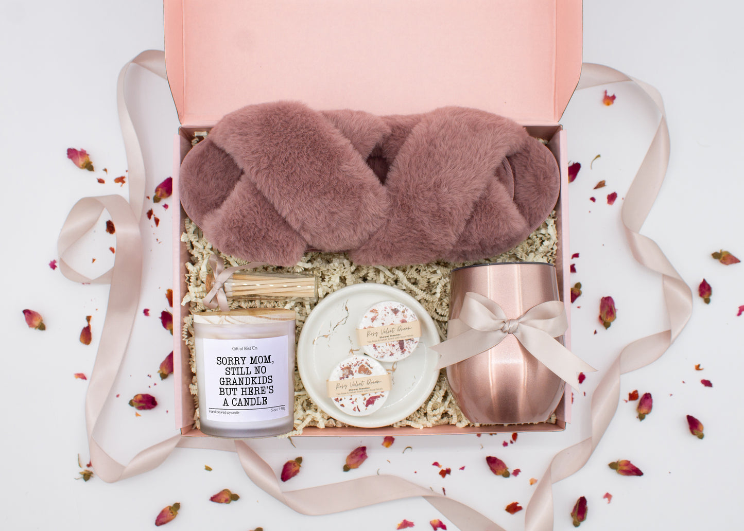 A luxury spa gift box for mom featuring a stainless steel tumbler, an aromatherapy candle with matches, therapeutic shower steamers, plush open-toe slippers, and gourmet caramel treats. All items are beautifully arranged for a pampering experience.