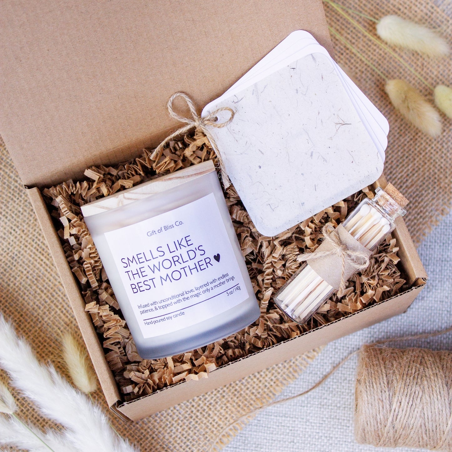 Candle gift box featuring a “Smells Like the World’s Best Mother” soy candle, glass match jar, and a rustic-style personalized message booklet tied with jute twine. Arranged in a kraft box with crinkle paper filler and neutral tones, this cozy, handcrafted gift is perfect for Mother’s Day or mom appreciation. A heartfelt and aesthetic set that combines warmth, functionality, and rustic charm.