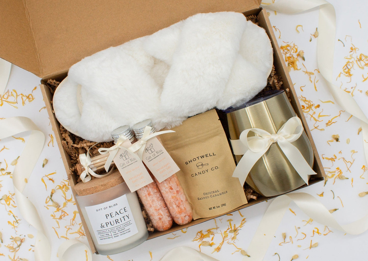 Luxury gift box for her. Featuring a stainless steel tumbler, an aromatherapy candle with matches, therapeutic bath salt, plush open-toe slippers, and gourmet caramel treats. All items are beautifully arranged for a pampering experience.