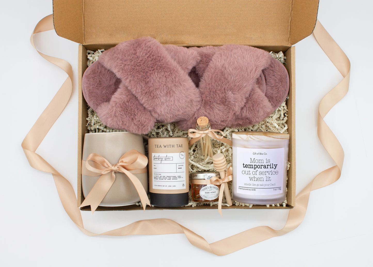Gift box in a premium Mauve theme featuring cozy mauve slippers, a minimalist ceramic mug tied with a champagne ribbon, Bombay Chai tea, a glass jar of honey with a wooden dipper, a white-tipped match jar, and a mother-themed labeled aromatherapy candle in a frosted jar. All items are nestled in ivory crinkle paper and elegantly arranged for a luxurious and thoughtful gift for moms.