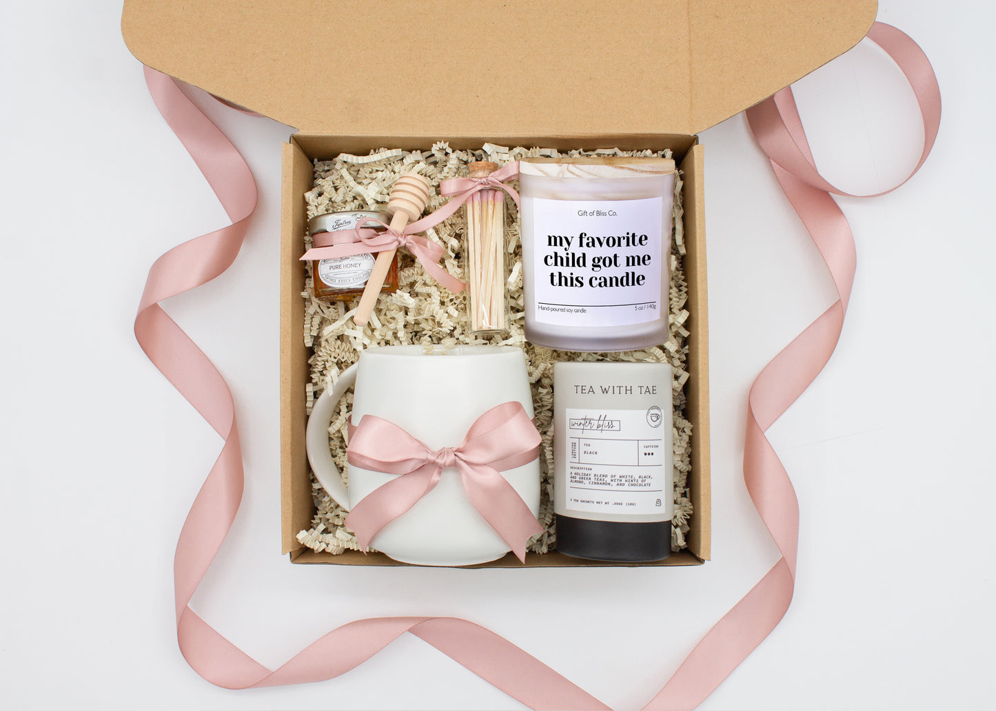 Gift box in a dusty pink theme featuring a white, minimalist ceramic mug tied with a dusty pink ribbon, Winter Bliss tea, a jar of pure honey with a wooden dipper, a white-tipped match jar, and a mother-themed labeled aromatherapy candle in a frosted glass jar. All items are nestled in ivory crinkle paper, making this a cozy and thoughtful gift for moms.