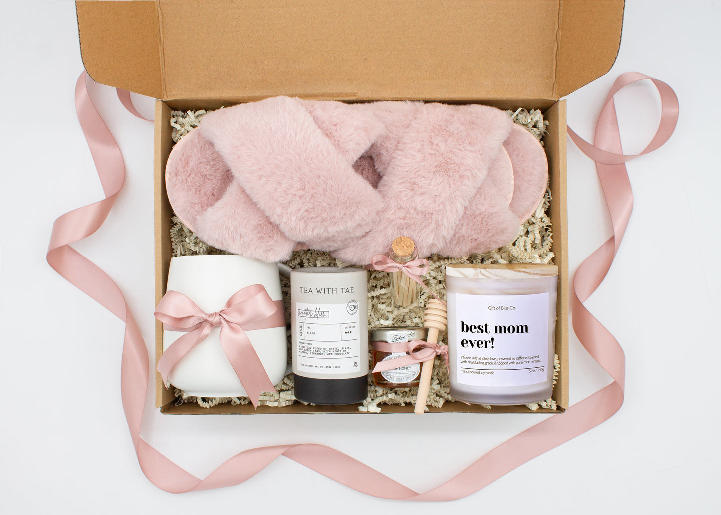 Gift box in a premium dusty pink theme featuring cozy dusty pink slippers, a minimalist ceramic mug tied with a dusty pink ribbon, Winter Bliss tea, a glass jar of honey with a wooden dipper, a white-tipped match jar, and a mother-themed labeled aromatherapy candle in a frosted jar. All items are nestled in ivory crinkle paper and elegantly arranged for a luxurious and thoughtful gift for moms.