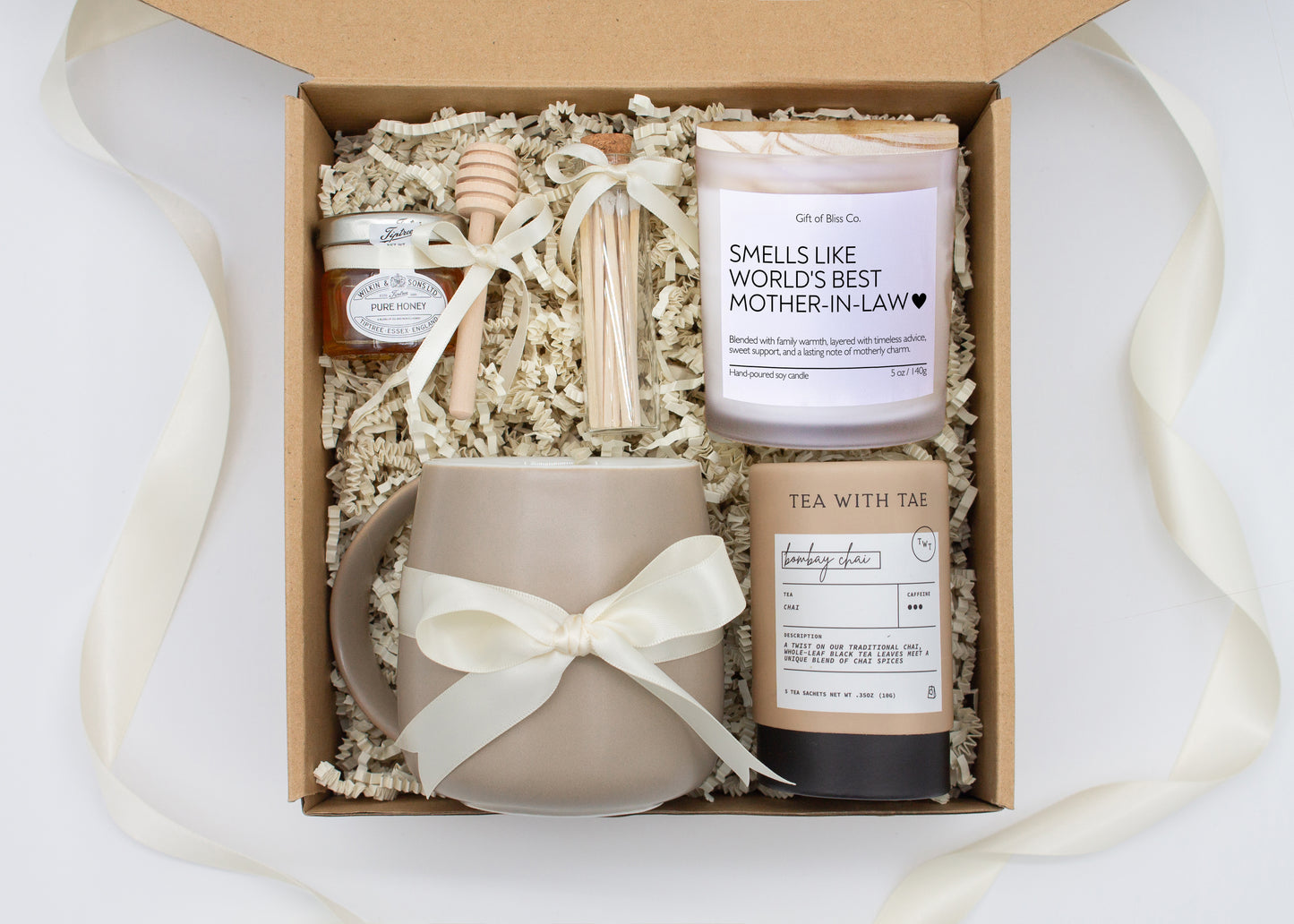 Gift box in an ivory theme featuring a taupe, minimalist ceramic mug tied with an ivory ribbon, Bombay Chai tea, a jar of pure honey with a wooden dipper, a white-tipped match jar, and a “Smells Like World's Best Mother-in-Law” labeled aromatherapy candle in a frosted glass jar. All items are nestled in ivory crinkle paper, making this a cozy and thoughtful gift for moms.