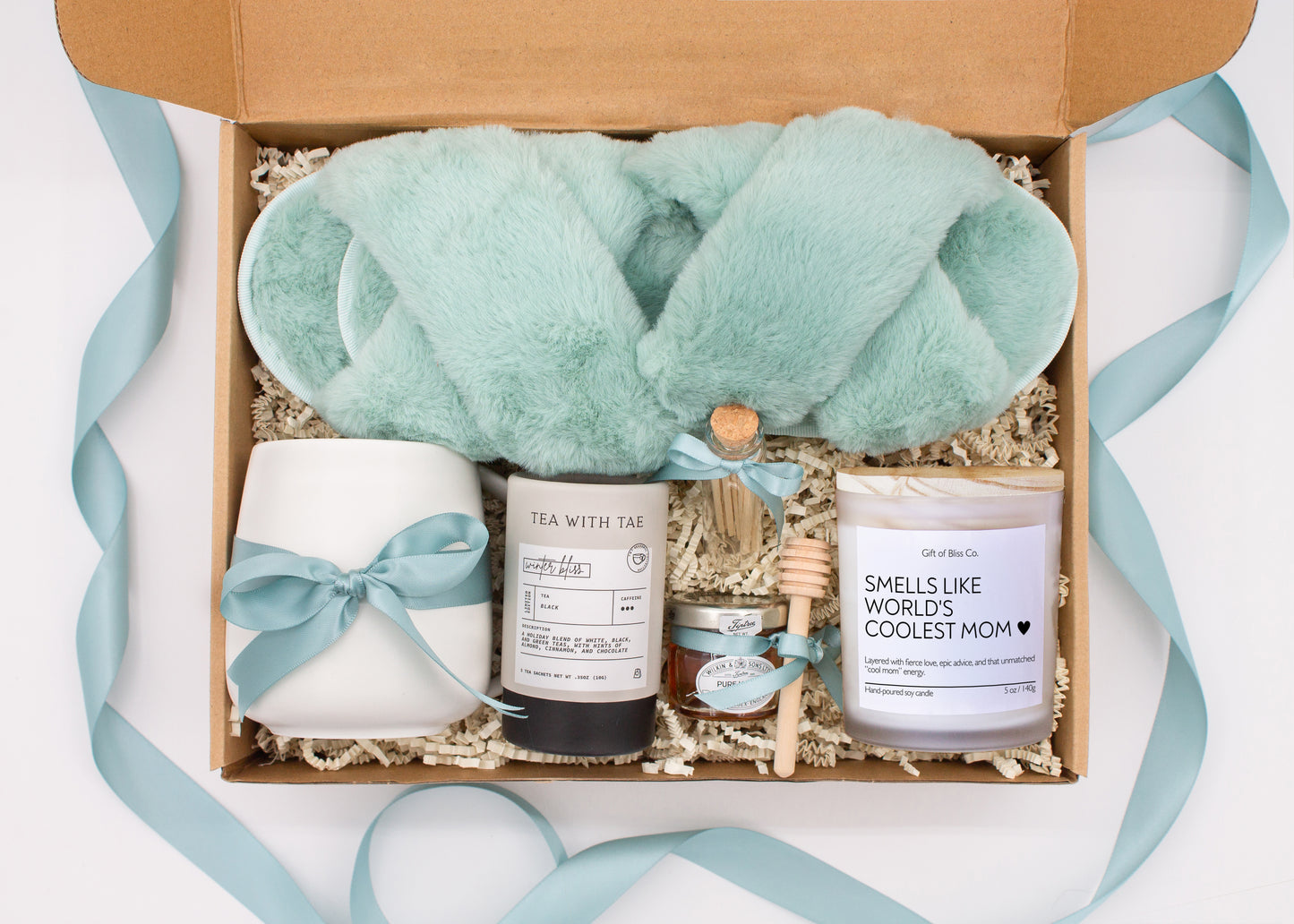 Gift box in a premium teal blue theme featuring cozy teal blue slippers, a minimalist ceramic mug tied with a teal blue ribbon, Winter Bliss tea, a glass jar of honey with a wooden dipper, a white-tipped match jar, and a mother-themed labeled aromatherapy candle in a frosted jar. All items are nestled in ivory crinkle paper and elegantly arranged for a luxurious and thoughtful gift for moms.