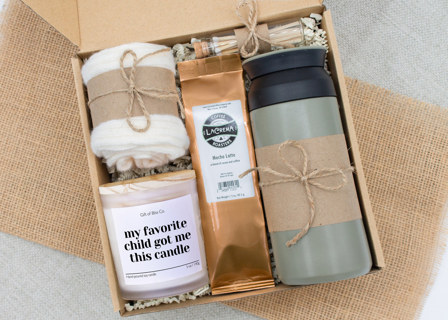 Rustic Retreat gift box in Basic Rustic Olive theme, featuring an olive color 12oz stainless steel tumbler, “my favorite child got me this candle" candle, safety match jar, mocha latte coffee, salted caramels, and cozy ivory socks. All items are arranged in kraft paper with jute cord for a charming, earthy presentation. A thoughtful, cozy gift set ideal for Mother’s Day or self-care.