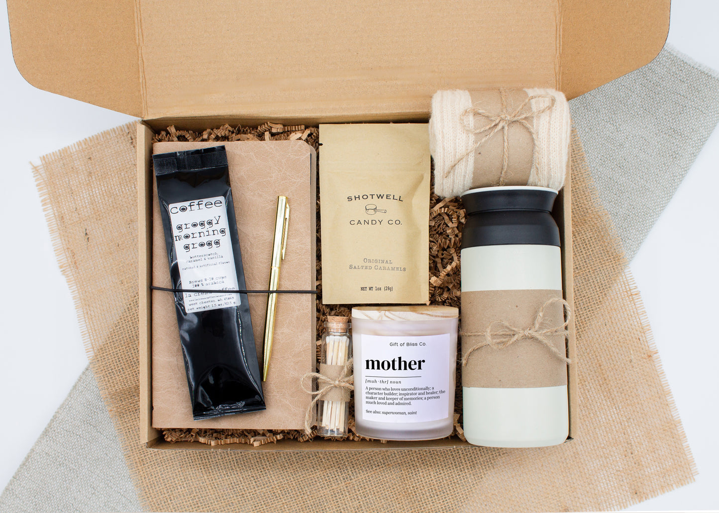 Rustic Retreat gift box in Premium Rustic Neutral theme, featuring a white 12oz stainless steel tumbler, “Mother" definition candle, safety match jar, mocha latte coffee, salted caramels, cozy ivory socks, vegan leather journal, and gold pen. All items are arranged in kraft paper with jute cord for a charming, earthy presentation. A thoughtful, cozy gift set ideal for Mother’s Day or self-care.
