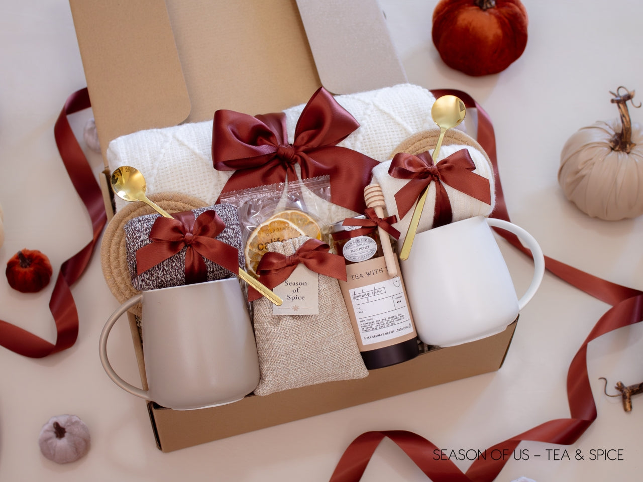 Gift box with artisan tea, pure honey, wooden dipper, dried orange slises, aromatic spice bag for simmer pots or mulling, 2 minimalist mugs, 2 long golden spoons, 2 pairs of cozy socks, soft white blanket, and 2 cotton yarn coasters, all decorated with satin ribbon in fall color, neatly arranged in a kraft gift box — a fall-inspired couples’ gift.