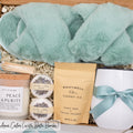 Luxury gift box for her. Featuring a stainless steel tumbler, an aromatherapy candle with matches, luxury bath bombs, plush open-toe slippers, and gourmet caramel treats. All items are beautifully arranged for a pampering experience.