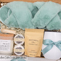 Luxury gift box for her. Featuring a stainless steel tumbler, an aromatherapy candle with matches, luxury bath bombs, plush open-toe slippers, and gourmet caramel treats. All items are beautifully arranged for a pampering experience.