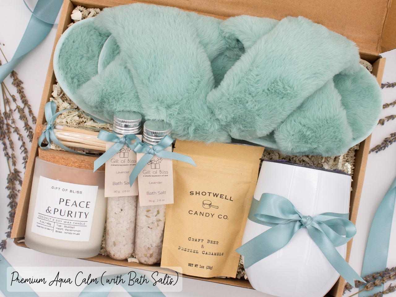 Luxury gift box for her. Featuring a stainless steel tumbler, an aromatherapy candle with matches, therapeutic bath salts, plush open-toe slippers, and gourmet caramel treats. All items are beautifully arranged for a pampering experience.