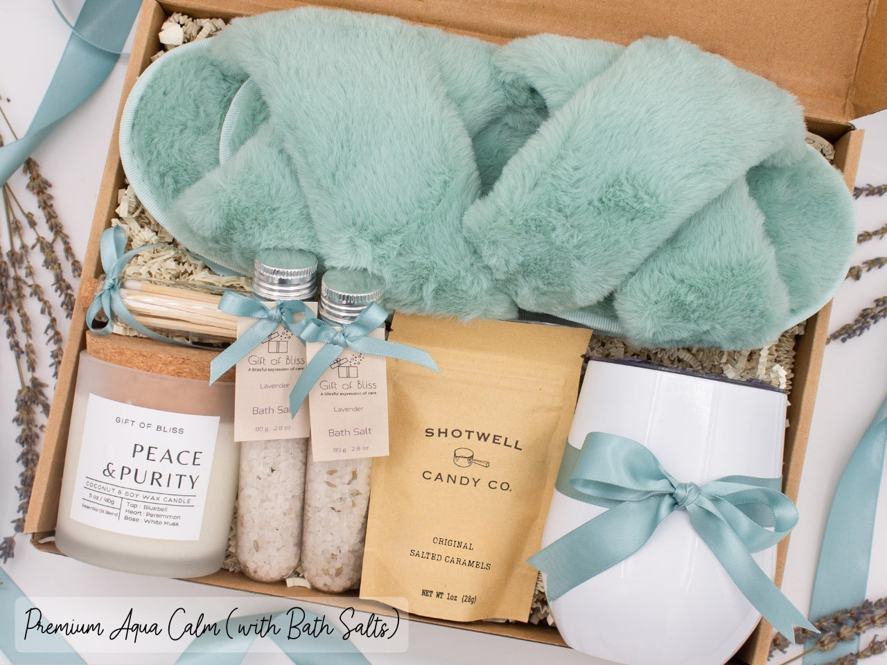 Luxury gift box for her. Featuring a stainless steel tumbler, an aromatherapy candle with matches, therapeutic bath salts, plush open-toe slippers, and gourmet caramel treats. All items are beautifully arranged for a pampering experience.