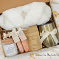 Luxury gift box for her. Featuring a stainless steel tumbler, an aromatherapy candle with matches, therapeutic bath salts, plush open-toe slippers, and gourmet caramel treats. All items are beautifully arranged for a pampering experience.