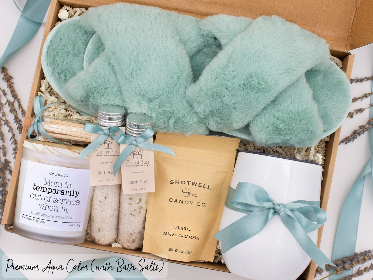 Luxury gift box for mom. Featuring a stainless steel tumbler, an aromatherapy candle with mom-themed label and charming match jar, therapeutic bath salts, plush open-toe slippers, and gourmet caramel treats. All items are beautifully arranged for a pampering experience.