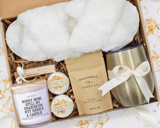 A luxury spa gift box for mom featuring a stainless steel tumbler, an aromatherapy candle with matches, therapeutic bath bombs, plush open-toe slippers, and gourmet caramel treats. All items are beautifully arranged for a pampering experience.
