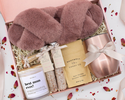 A luxury spa gift box for mom featuring a stainless steel tumbler, an aromatherapy candle with matches, therapeutic bath salt, plush open-toe slippers, and gourmet caramel treats. All items are beautifully arranged for a pampering experience.