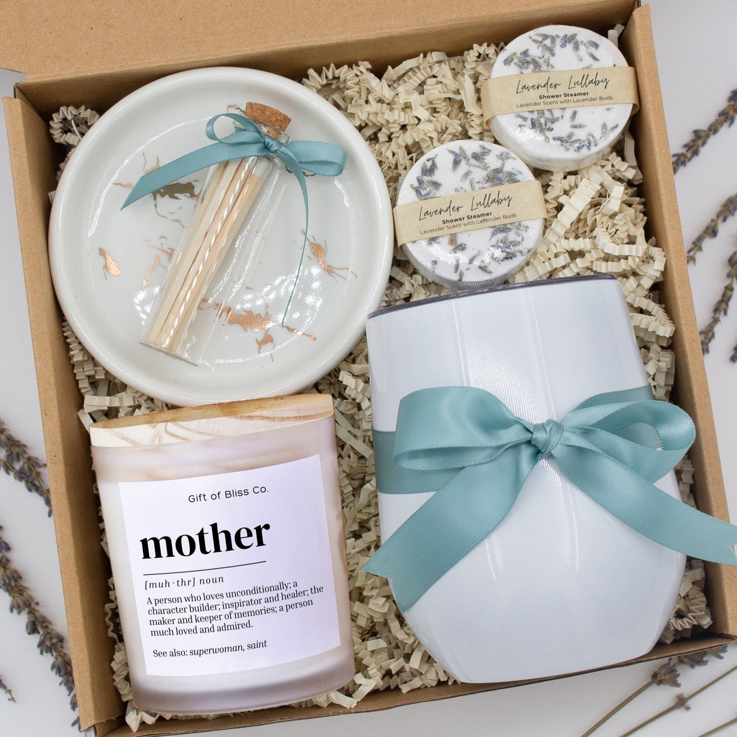 A luxury spa gift box for mom featuring a stainless steel tumbler, an aromatherapy candle with matches, therapeutic shower steamers, and gourmet caramel treats. All items are beautifully arranged for a pampering experience.