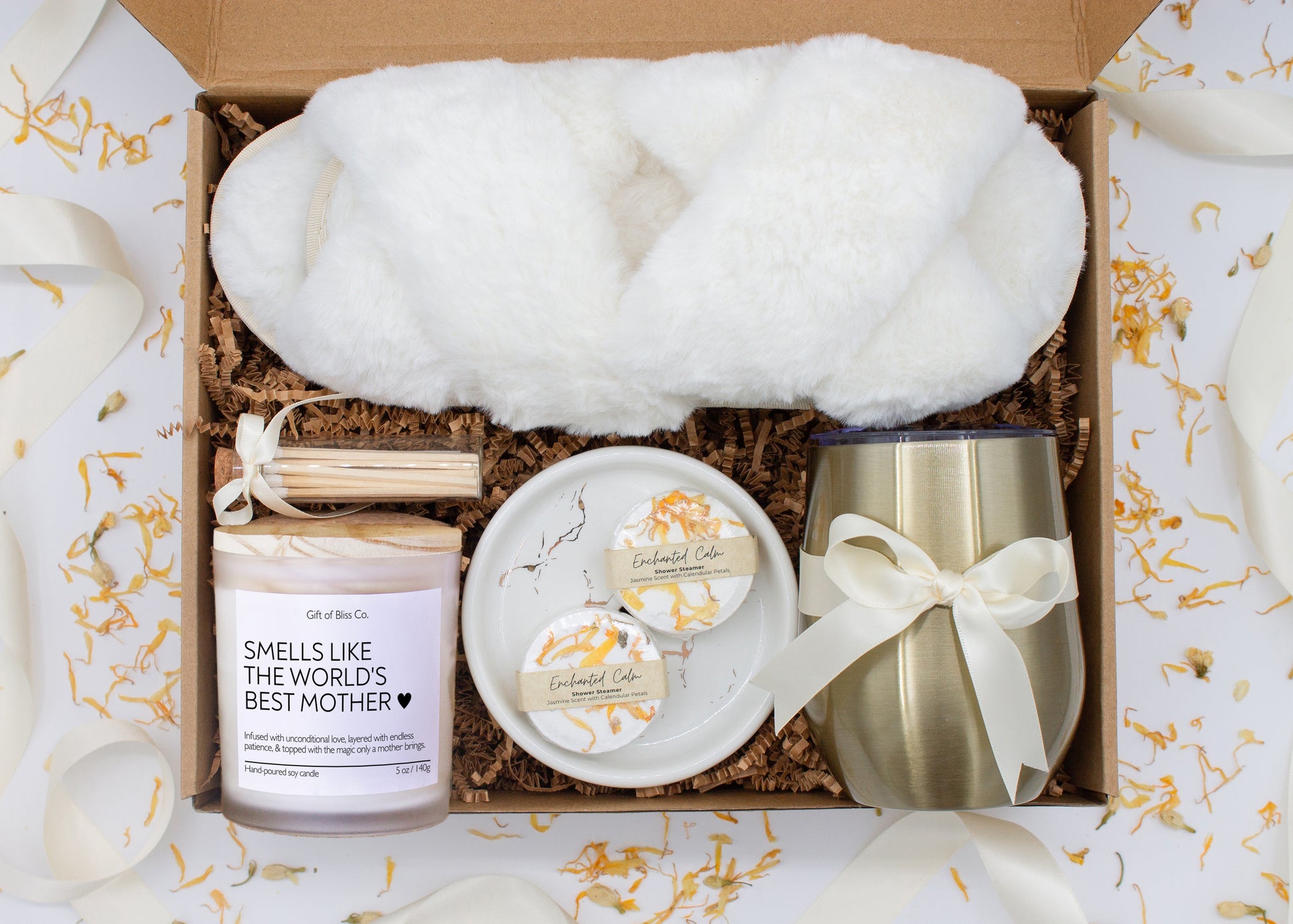 A luxury spa gift box for mom featuring a stainless steel tumbler, an aromatherapy candle with matches, therapeutic shower steamers, plush open-toe slippers, and gourmet caramel treats. All items are beautifully arranged for a pampering experience.