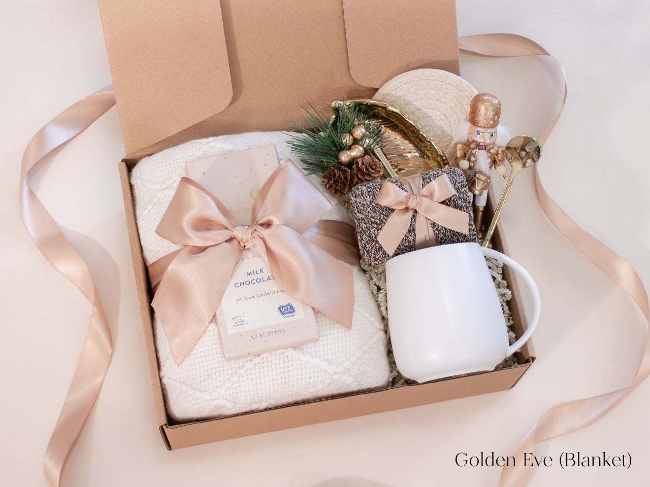 Elegant champagne-toned holiday gift box for him featuring a white mug with gold spoon, cozy knit socks, artisan chocolate bar, and soft ivory blanket tied with a satin ribbon - accented with a nutcracker figurine and gold leaf dish for a refined, festive touch.