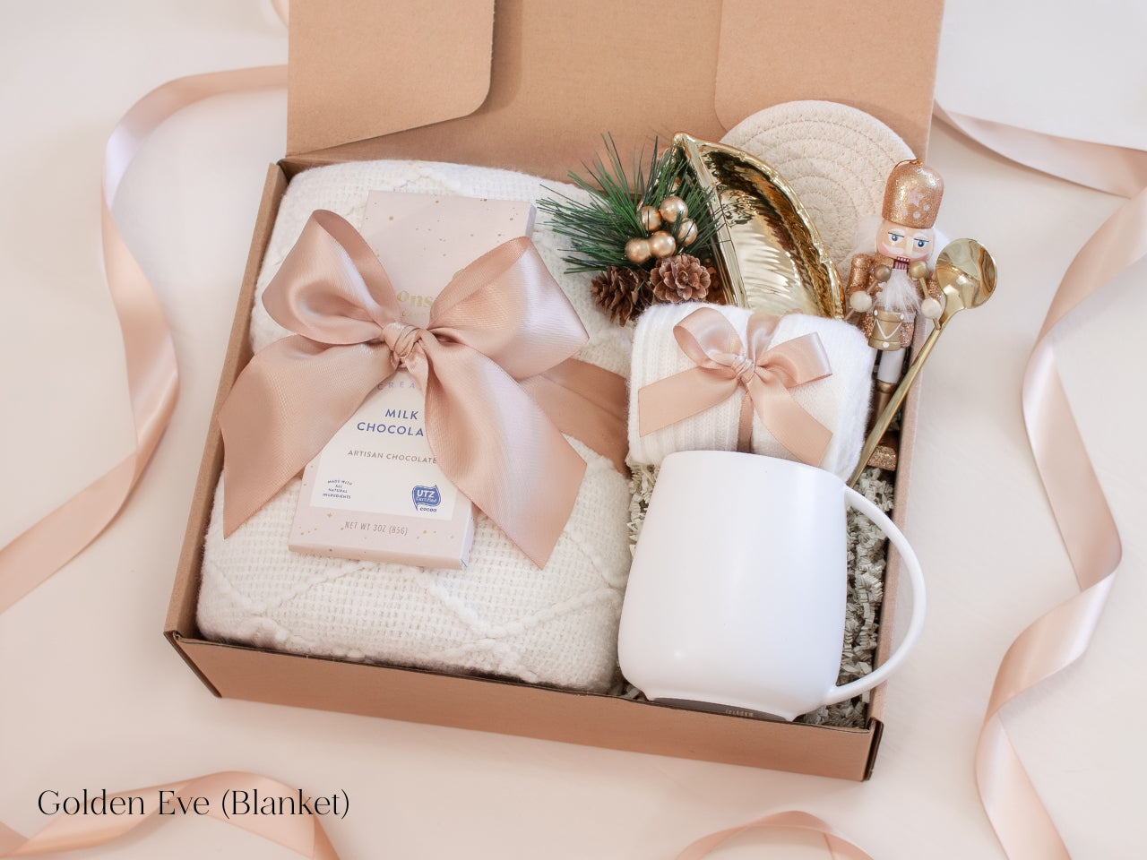 Elegant champagne-toned holiday gift box for him featuring a white mug with gold spoon, cozy knit socks, artisan chocolate bar, and soft ivory blanket tied with a satin ribbon - accented with a nutcracker figurine and gold leaf dish for a refined, festive touch.
