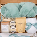 Luxury gift box for her. Featuring a stainless steel tumbler, an aromatherapy candle with matches, luxury bath bombs, plush open-toe slippers, and gourmet caramel treats. All items are beautifully arranged for a pampering experience.