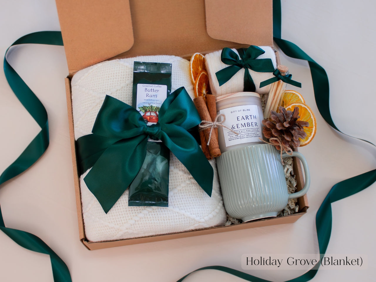Rustic green holiday gift box for him featuring a ribbed ceramic mug, Earth & Ember candle, butter rum coffee, cinnamon sticks, dried orange slices, pinecone, and a soft white blanket - finished with deep green ribbons for a cozy, nature-inspired winter aesthetic.