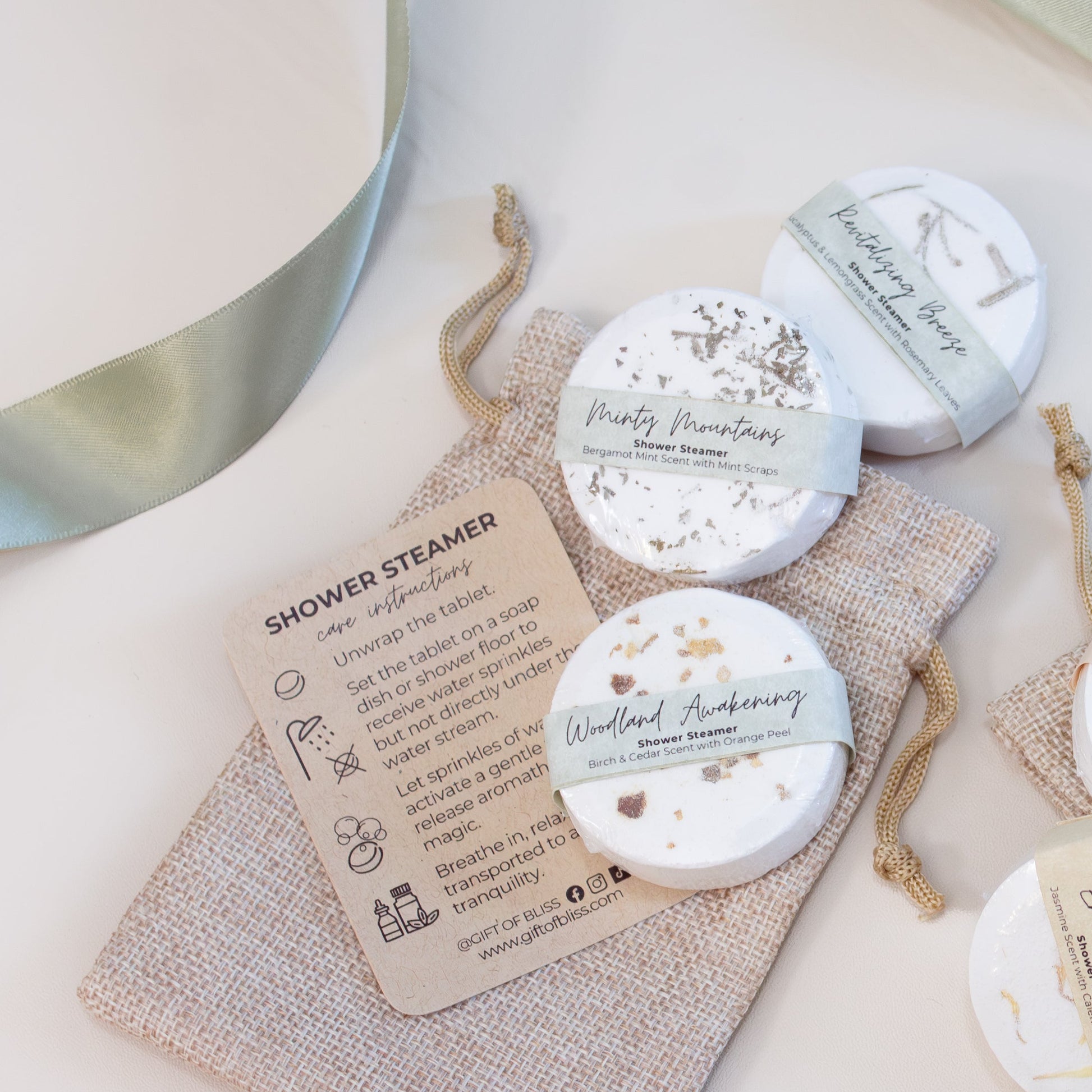 A flat-lay photo of three white shower steamers, each labeled with soft pastel bands and natural names like “Minty Mountains,” “Woodland Awakening,” and “Revitalizing Breeze.” The steamers are arranged beside a small burlap drawstring bag and a printed instruction card, with a sage green satin ribbon woven throughout the scene. The overall look feels clean, calm, and spa-inspired — perfect for a self-care or relaxation gift set.