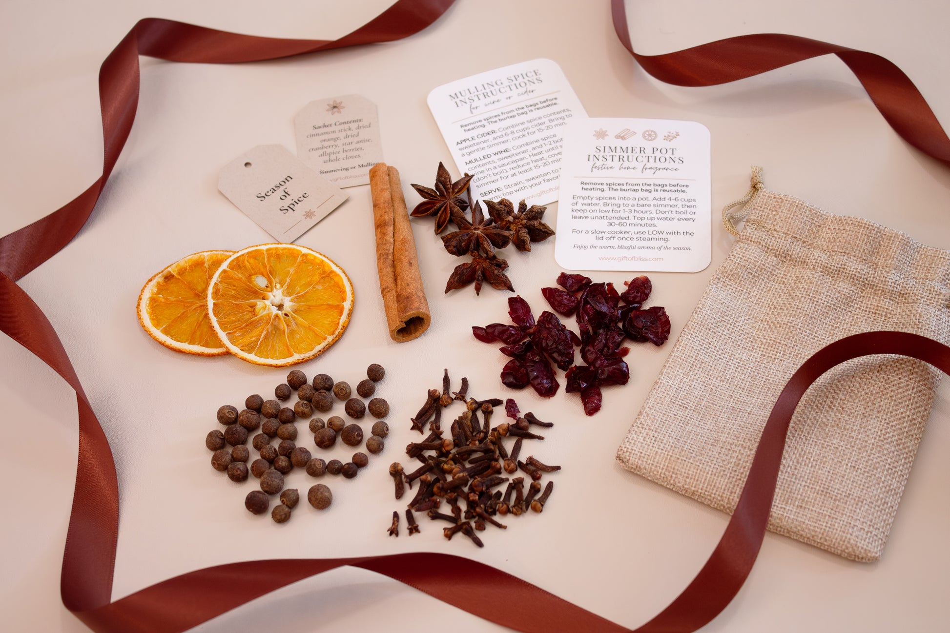 Flat lay of an aromatic spice kit for simmer pots and mulled drinks, featuring dried orange slices, cinnamon stick, star anise, cloves, cranberries, allspice, and sachet bags, arranged with instruction cards and a reusable burlap pouch, accented by a burgundy ribbon.