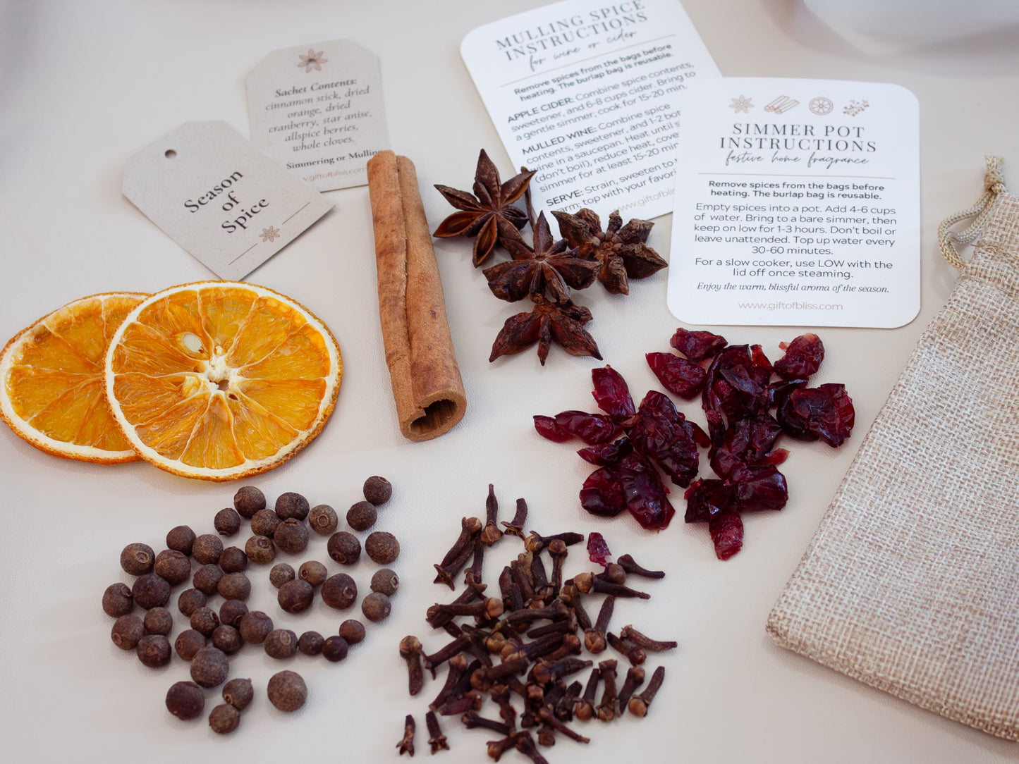 Flat lay of an aromatic spice kit for simmer pots and mulled drinks, featuring dried orange slices, cinnamon stick, star anise, cloves, cranberries, allspice, and sachet bags, arranged with instruction cards and a reusable burlap pouch.