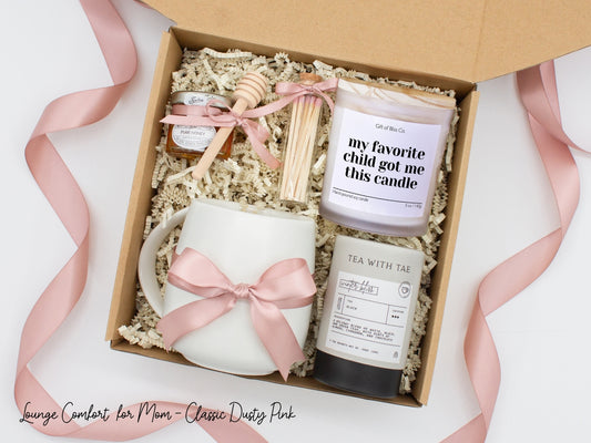 Gift box in a basic dusty pink theme featuring a white, minimalist ceramic mug tied with a dusty pink ribbon, Winter Bliss tea, a jar of pure honey with a wooden dipper, a white-tipped match jar, and a mother-themed labeled aromatherapy candle in a frosted glass jar. All items are nestled in ivory crinkle paper, making this a cozy and thoughtful gift for moms.