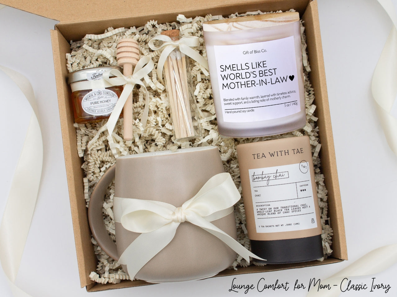 Gift box in a basic ivory theme featuring a taupe, minimalist ceramic mug tied with an ivory ribbon, Bombay Chai tea, a jar of pure honey with a wooden dipper, a white-tipped match jar, and a “Smells Like World's Best Mother-in-Law” labeled aromatherapy candle in a frosted glass jar. All items are nestled in ivory crinkle paper, making this a cozy and thoughtful gift for moms.