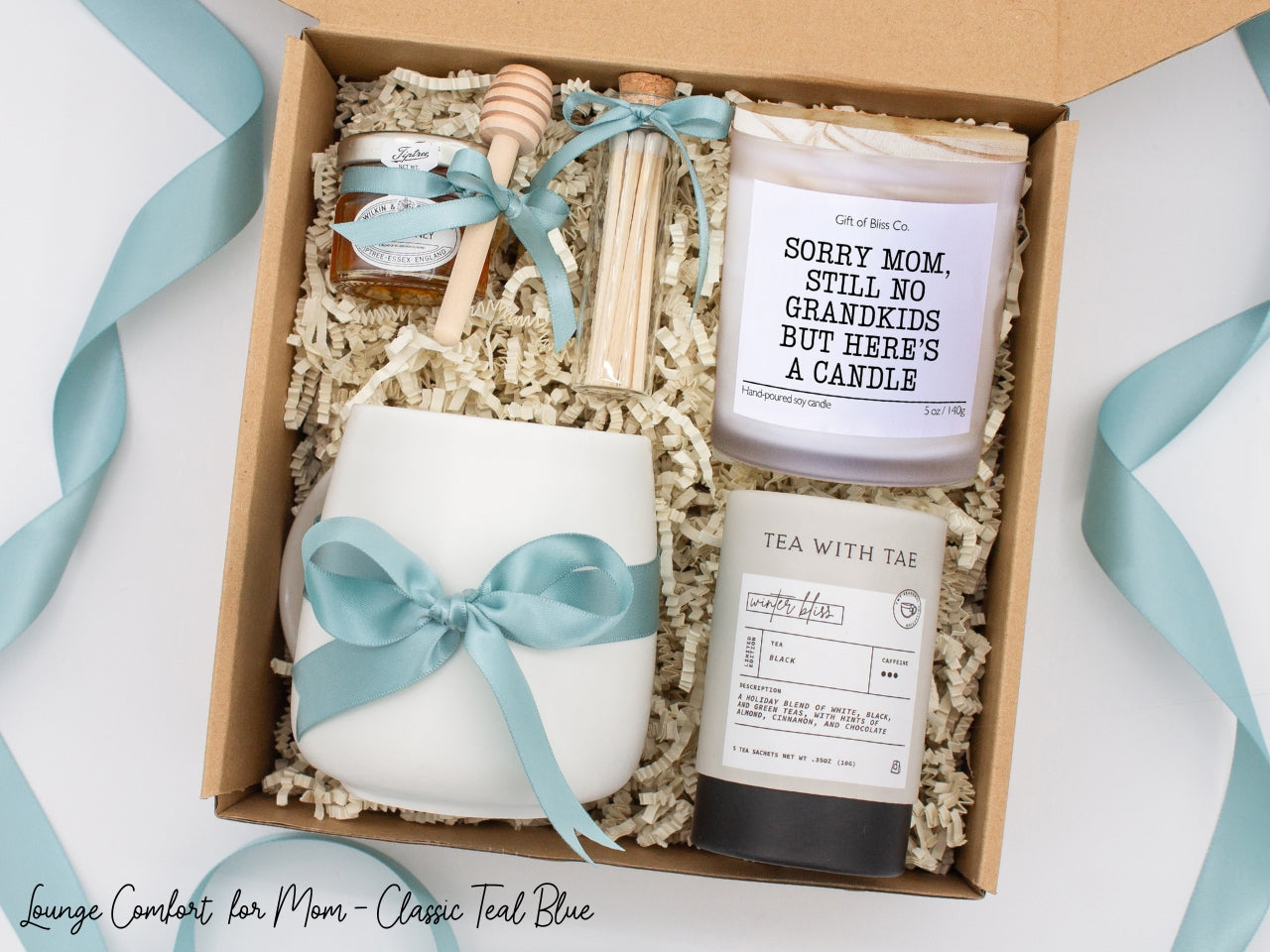 Gift box in a basic teal blue theme featuring a white, minimalist ceramic mug tied with a teal blue ribbon, Winter Bliss tea, a jar of pure honey with a wooden dipper, a white-tipped match jar, and a mother-themed labeled aromatherapy candle in a frosted glass jar. All items are nestled in ivory crinkle paper, making this a cozy and thoughtful gift for moms.