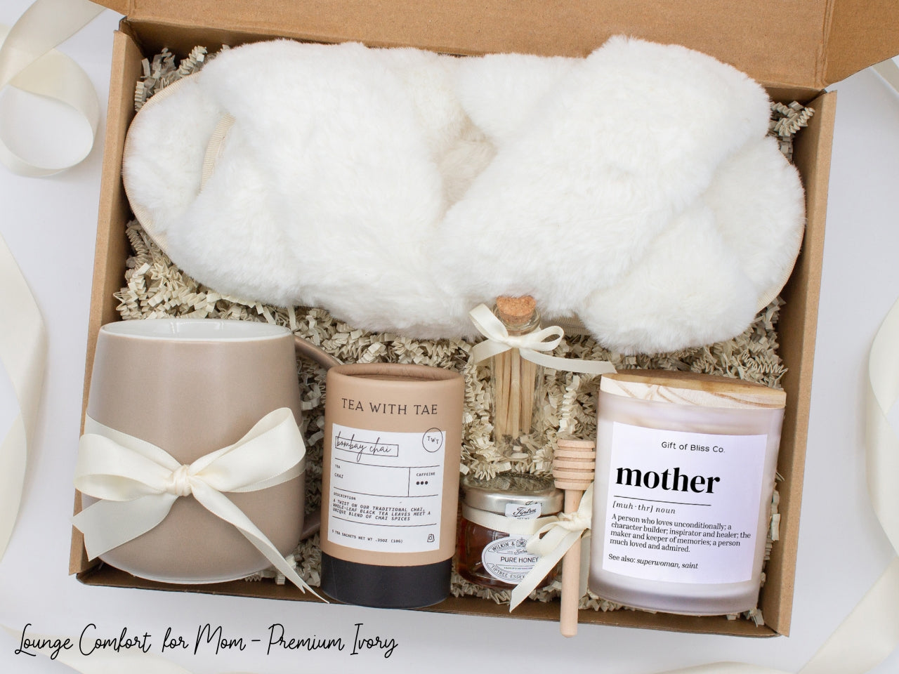 Gift box in a premium ivory theme featuring cozy white slippers, a minimalist ceramic mug tied with an ivory ribbon, Bombay Chai tea, a glass jar of honey with a wooden dipper, a white-tipped match jar, and a "mother” definition labeled aromatherapy candle in a frosted jar. All items are nestled in ivory crinkle paper and elegantly arranged for a luxurious and thoughtful gift for moms.