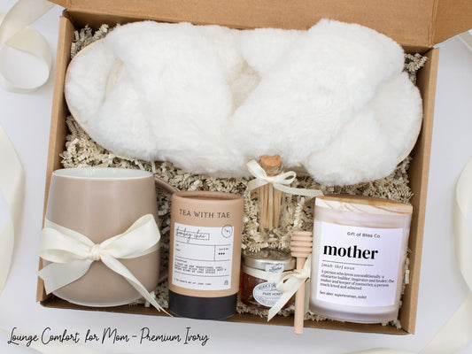 Gift box in a premium ivory theme featuring cozy white slippers, a minimalist ceramic mug tied with an ivory ribbon, Bombay Chai tea, a glass jar of honey with a wooden dipper, a white-tipped match jar, and a "mother” definition labeled aromatherapy candle in a frosted jar. All items are nestled in ivory crinkle paper and elegantly arranged for a luxurious and thoughtful gift for moms.