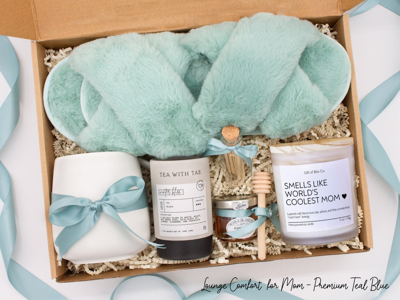 Gift box in a premium teal blue theme featuring cozy teal blue slippers, a minimalist ceramic mug tied with a teal blue ribbon, Winter Bliss tea, a glass jar of honey with a wooden dipper, a white-tipped match jar, and a mother-themed labeled aromatherapy candle in a frosted jar. All items are nestled in ivory crinkle paper and elegantly arranged for a luxurious and thoughtful gift for moms.