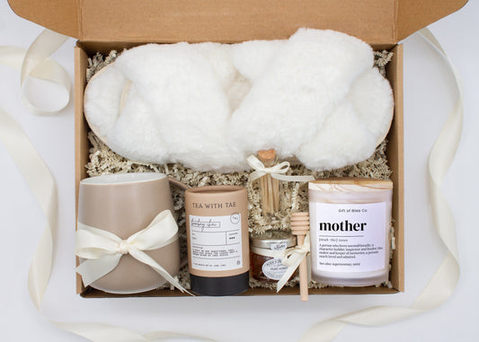 Gift box in a premium ivory theme featuring cozy white slippers, a minimalist ceramic mug tied with an ivory ribbon, Bombay Chai tea, a glass jar of honey with a wooden dipper, a white-tipped match jar, and a "mother” definition labeled aromatherapy candle in a frosted jar. All items are nestled in ivory crinkle paper and elegantly arranged for a luxurious and thoughtful gift for moms.