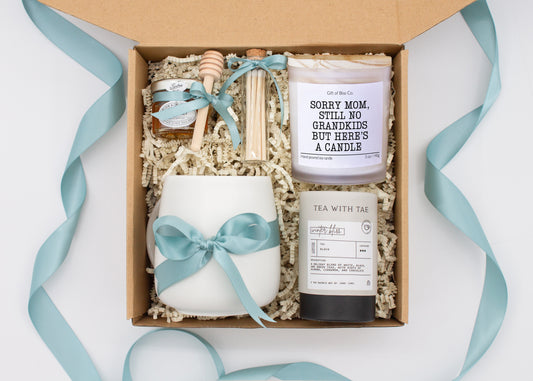 Gift box in a teal blue theme featuring a white, minimalist ceramic mug tied with a teal blue ribbon, Winter Bliss tea, a jar of pure honey with a wooden dipper, a white-tipped match jar, and a mother-themed labeled aromatherapy candle in a frosted glass jar. All items are nestled in ivory crinkle paper, making this a cozy and thoughtful gift for moms.