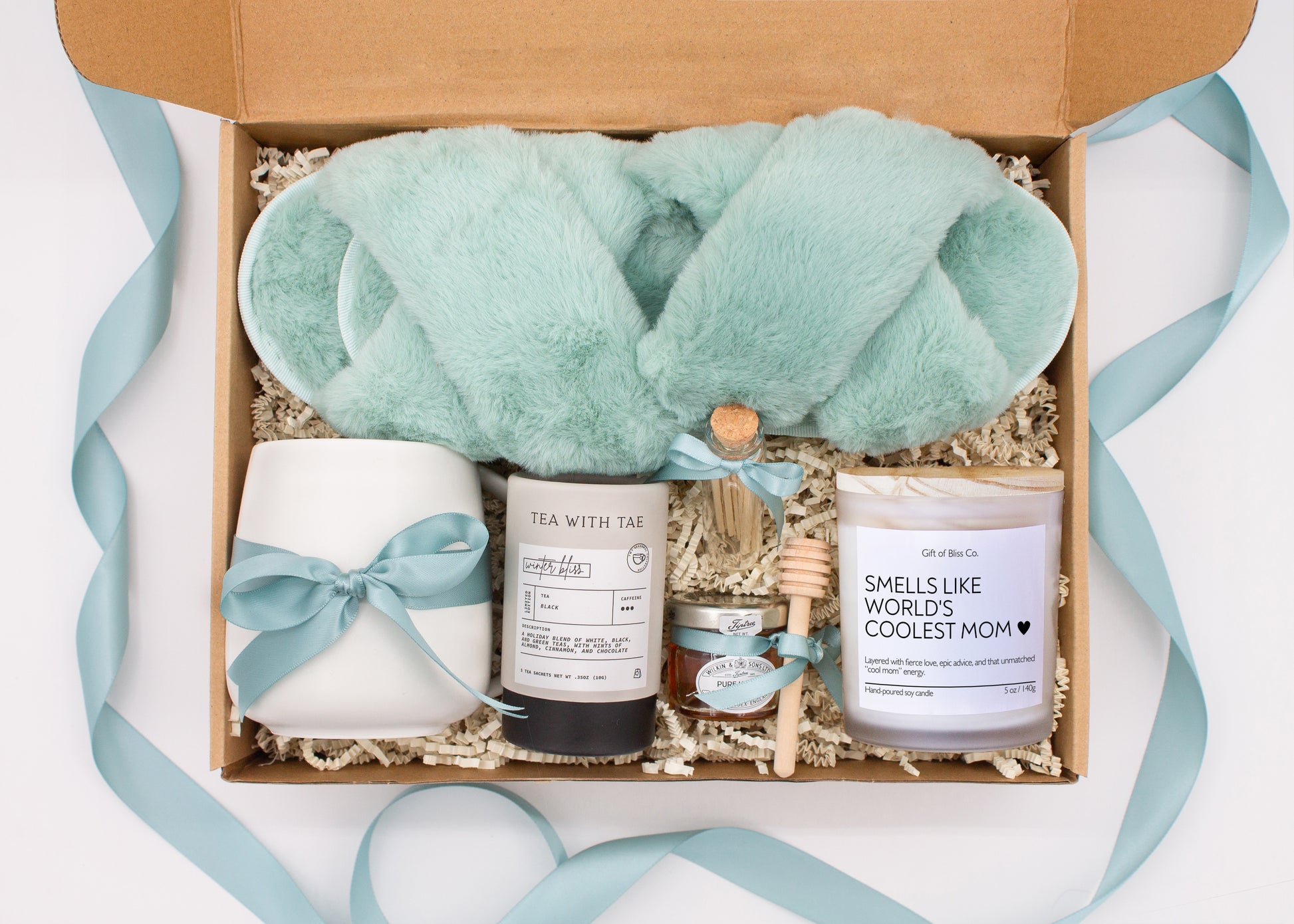 Gift box in a premium teal blue theme featuring cozy teal blue slippers, a minimalist ceramic mug tied with a teal blue ribbon, Winter Bliss tea, a glass jar of honey with a wooden dipper, a white-tipped match jar, and a mother-themed labeled aromatherapy candle in a frosted jar. All items are nestled in ivory crinkle paper and elegantly arranged for a luxurious and thoughtful gift for moms.
