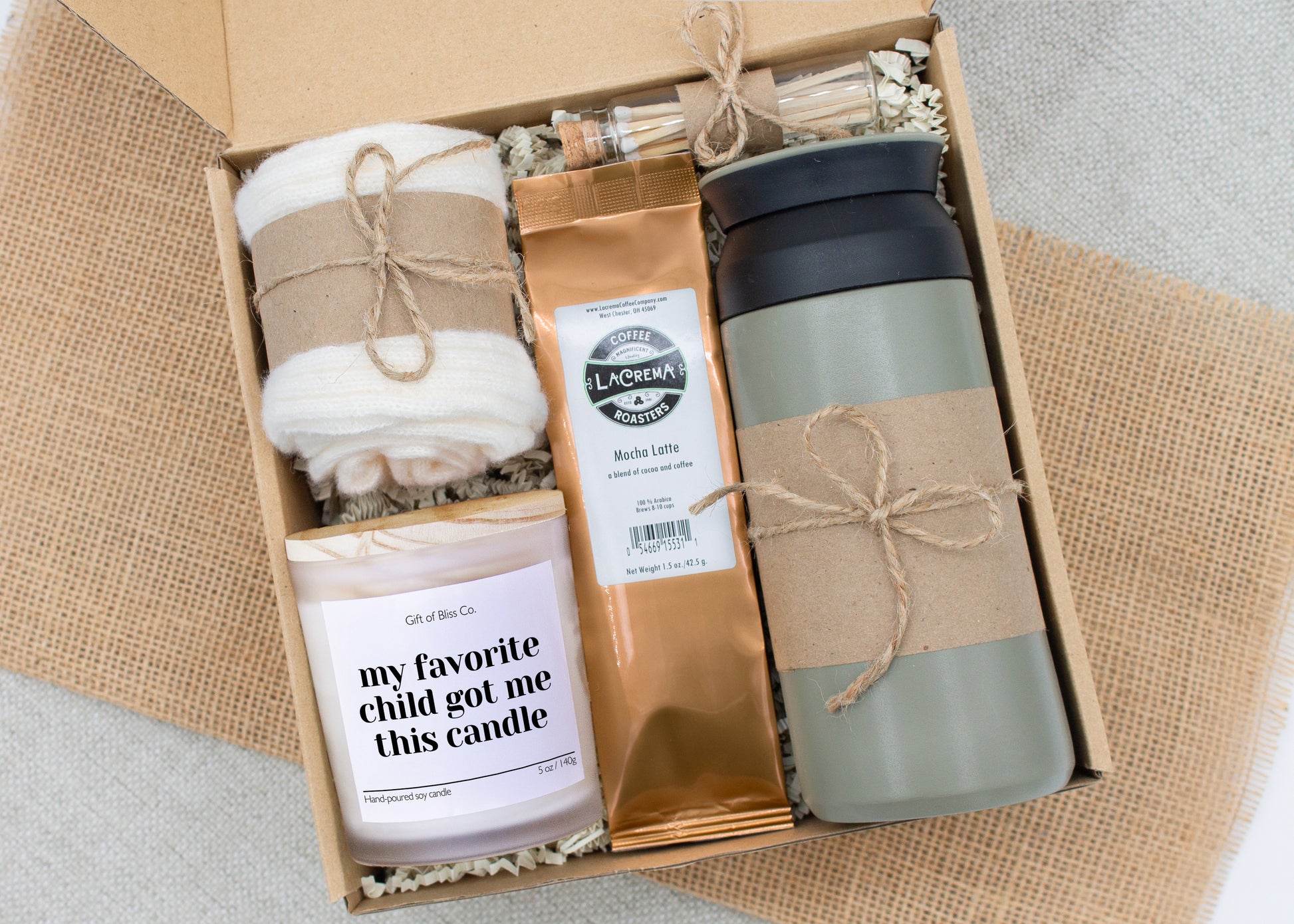 Rustic Retreat gift box in Basic Rustic Olive theme, featuring an olive color 12oz stainless steel tumbler, “my favorite child got me this candle" candle, safety match jar, mocha latte coffee, salted caramels, and cozy ivory socks. All items are arranged in kraft paper with jute cord for a charming, earthy presentation. A thoughtful, cozy gift set ideal for Mother’s Day or self-care.