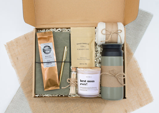 Rustic Retreat gift box in Premium Rustic Olive theme, featuring an olive color 12oz stainless steel tumbler, “best mom ever” candle, safety match jar, mocha latte coffee, salted caramels, cozy ivory socks, vegan leather journal, and gold pen. All items are arranged in kraft paper with jute cord for a charming, earthy presentation. A thoughtful, cozy gift set ideal for Mother’s Day or self-care.