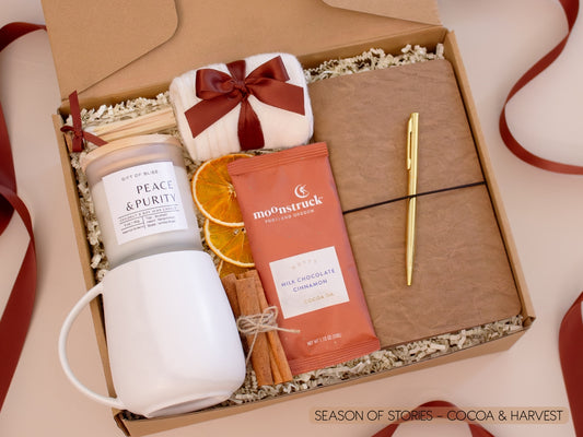 A cozy kraft gift box filled with a white ceramic mug, Earth & Ember candle, soft socks, rich Moonstruck cocoa chocolate, cinnamon sticks, and dried orange slices, alongside a vegan leather notebook and gold pen—perfect for quiet moments of writing and sipping.