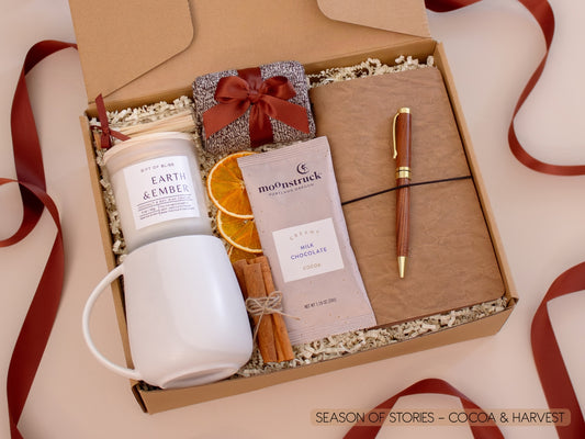 A cozy kraft gift box filled with a white ceramic mug, Earth & Ember candle, soft socks, rich Moonstruck cocoa chocolate, cinnamon sticks, and dried orange slices, alongside a vegan leather notebook and gold pen—perfect for quiet moments of writing and sipping.