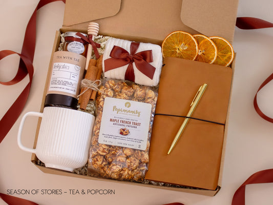 A heartwarming gift box with a ribbed white mug, Bombay Chai tea and honey with wooden dipper, maple French toast gourmet popcorn, a vegan leather notebook with gold pen, soft socks, cinnamon sticks, and dried orange slices—made for sharing stories over tea and treats.