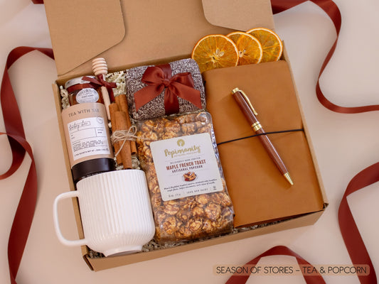 A heartwarming gift box with a ribbed white mug, Bombay Chai tea and honey with wooden dipper, maple French toast gourmet popcorn, a vegan leather notebook with gold pen, soft socks, cinnamon sticks, and dried orange slices—made for sharing stories over tea and treats.